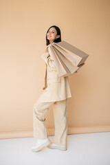 full length of cheerful asian woman in stylish pantsuit posing with shopping bags on beige...
