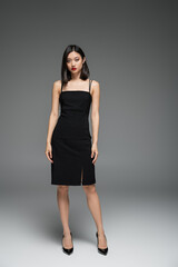 full length of slender asian woman in black strap dress looking at camera on grey background.