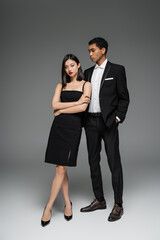 full length of asian woman in black dress and african american guy in elegant suit posing on grey background.