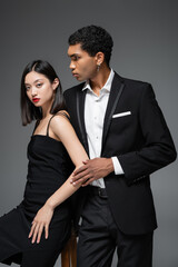 african american man in black blazer looking away near asian woman in elegant strap dress isolated on grey.