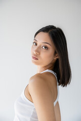 charming asian woman with perfect skin and brunette hair looking at camera isolated on grey.