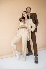 full length of african american man hugging trendy asian model in ivory pantsuit on beige background.