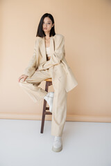 brunette asian woman in stylish pantsuit and boots sitting on stool on beige background.