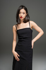 brunette asian woman in black stylish dress posing with hand on hip on grey background.