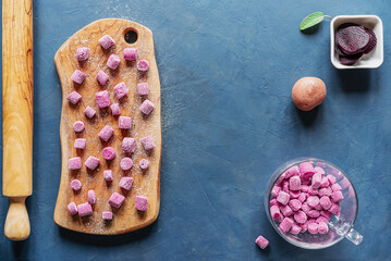 Beetroot and potato homemade gnocchi on wooden cutting board and in a glass mug, ingredients, sage leaf, rolling pin, dark blue background, top view