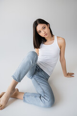 pretty asian woman in white tank top and jeans looking at camera while sitting on grey background.