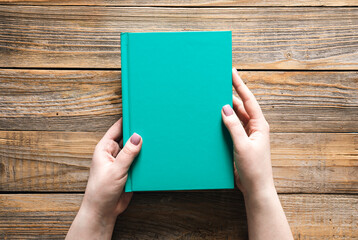 Closed green book in female hands on a wooden background, top view.