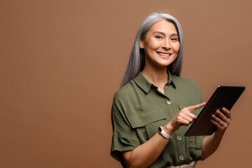 Mature 50s asian woman smiling and holding digital tablet, standing over brown background, japanese lady touching touch screen with index finger, looking at the camera standing isolated