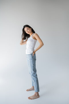full length of barefoot asian model in tank top posing with hand in back pocket of blue jeans on grey background.