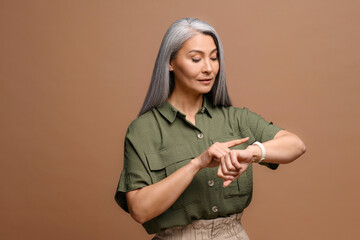 Punctual mature asian woman checking time on wrist watch isolated on brown background, responsible...