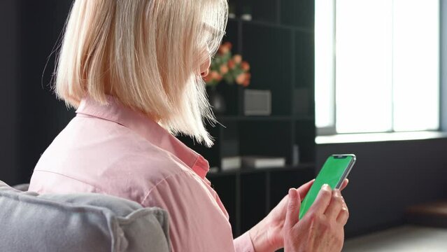 View from shoulder of caucasian blonde  woman doing swipe up gesture on chroma key of modern smartphone. Mature female resting at home and using cell phone.