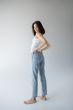 full length of barefoot asian woman in white tank top and blue jeans holding hands in back pockets on grey background.