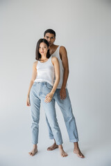 full length of young and barefoot interracial couple posing in white tank tops and blue jeans on grey background.