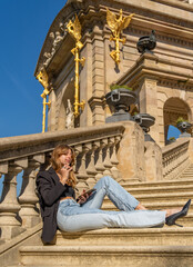 Fototapeta premium Young, tanned, beautiful woman in a black top and jeans with sunglasses and long hair, sitting on a granite staircase sideways with her legs stretched out looking, reading, typing on her mobile phone