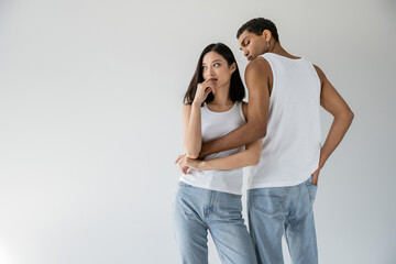 Naklejka premium african american man in white tank top and jeans embracing dreamy asian woman looking away isolated on grey.