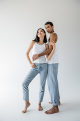 full length of african american man in tank top and jeans embracing cheerful asian model on grey background.