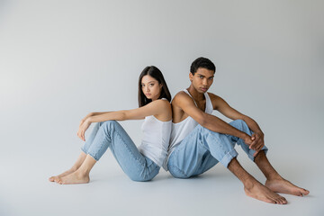 Fototapeta na wymiar full length of barefoot and stylish interracial couple in jeans and tank tops sitting back to back on grey background.
