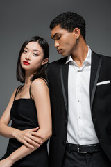 asian woman in black strap dress posing near young african american man in elegant blazer isolated on grey.
