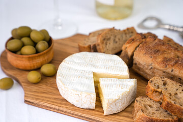 Camembert cheese, sliced fresh baguette, olives on a wooden board on the table. A wineglass and a...