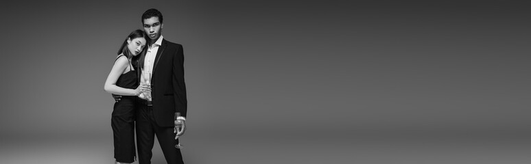 stylish multiethnic couple in black elegant attire holding champagne glasses isolated on grey, banner.