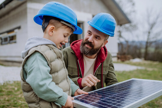 Father and his little son holding solar panel, in front of their new unfinished house.