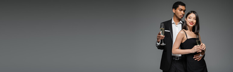 smiling asian woman and elegant african american man posing with champagne glasses isolated on grey, banner.