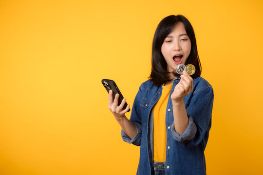 Happy young asian woman wearing yellow t-shirt denim shirt holding digital crypto currency coin and smartphone isolated on yellow background. Digital currency financial concept.