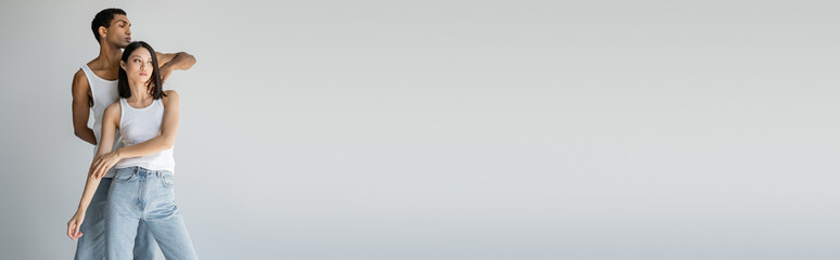 interracial couple in blue jeans and white tank tops posing isolated on grey, banner.