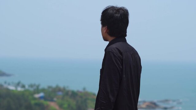 Indian man looking at the Vagator beach while sitting on the wall of the Chapora fort at Goa in India. Tourist enjoys the views of the beach from above the hill. Summer holiday background.