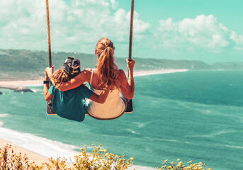 Mother and son having fun on swing above atlantic ocean- Portugal, Nazare- family love, vacation,...