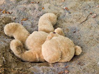 Light brown teddy bear laying down in the sandbox at childrens playground in summer.