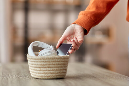 digital detox and technology concept - close up of hand holding smartphone and different gadgets in wicker basket on table at home