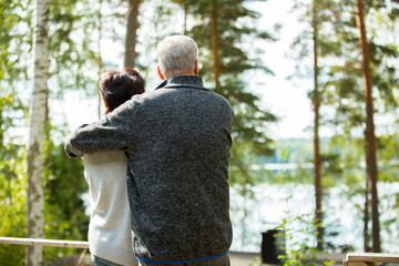 Mature couple standing on porch and hugging. They are smiling and looking at the beautiful view - forest and lake. Happy senior couple embracing each other on the wooden terrace of the house. 