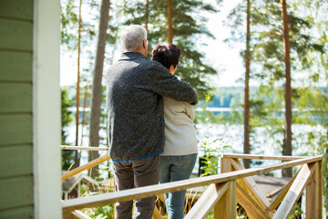 Mature couple standing on porch and hugging. They are smiling and looking at the beautiful view - forest and lake. Happy senior couple embracing each other on the wooden terrace of the house. 