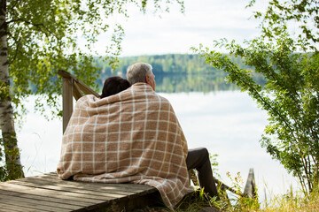 Mature couple sitting on wooden steps and hugging. They are wrapped in blanket, smiling and looking at beautiful view - forest and lake. Happy senior couple embracing each other 