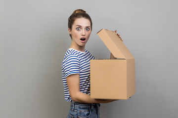 Side view portrait of surprised shocked woman wearing striped T-shirt standing with carton box and...
