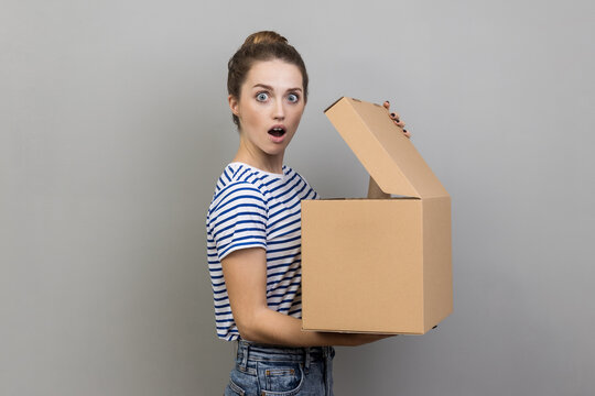Side view portrait of surprised shocked woman wearing striped T-shirt standing with carton box and looking at camera with big eyes and widely open mouth. Indoor studio shot isolated on gray background