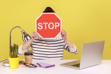 Woman office worker sitting at workplace hiding face behind red stop traffic sign avoiding...