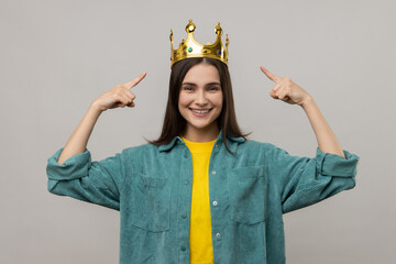 Happy woman pointing fingers on golden crown on her head, showing her leadership qualities, concept of authority, wearing casual style jacket. Indoor studio shot isolated on gray background.