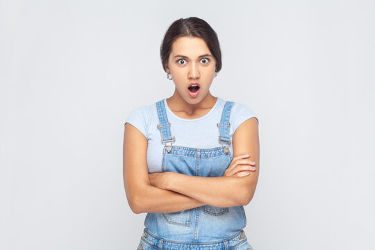 Portrait of astonished shoked woman wearing denim overalls having stunned and shocked look, standing with crossed arms and open mouth. Indoor studio shot isolated on gray background.