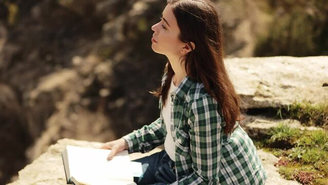 A long-haired girl reads a book in the sun. A young woman reads the Bible outdoors. A woman holds a Bible in her hands and studies the word of God on top of a mountain.