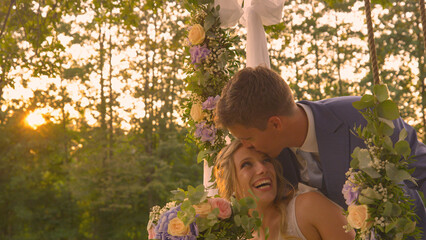 LENS FLARE: Handsome groom kisses his newlywed wife, who is sitting on a swing