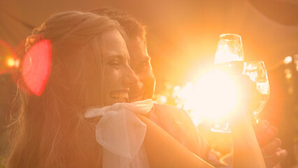 LENS FLARE, CLOSE UP: Smiling bride and groom with raised glasses of champagne