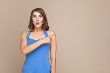 Obraz premium Portrait of surprised woman with wavy hair showing something aside at blank wall, being astonished, advertisement area, wearing blue dress. Indoor studio shot isolated on light brown background.