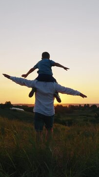 Vertical Screen: Dad walking in meadow with little son sitting on his shoulders, both spreading arms as if flying. Following shot family spending great time together. Concept of bonding