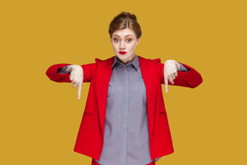 Portrait of strict serious woman with red lips standing pointing both index fingers down, saying here and right now, wearing red jacket. Indoor studio shot isolated on yellow background.