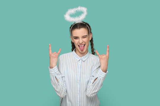 Portrait of extremely happy teenager girl with braids wearing striped shirt and nimb over her hair, showing rock and roll gesture, showing tongue. Indoor studio shot isolated on green background.