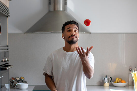 Serious ethnic man in earbuds throwing up tomato in kitchen
