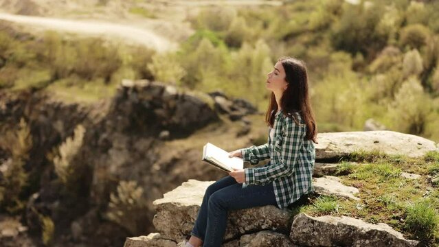 A beautiful young woman is sitting on the edge of a cliff and reading a book, enjoying the view and a sunny day. An attractive girl relaxes and studies outdoors with an incredible view. Slow motion 