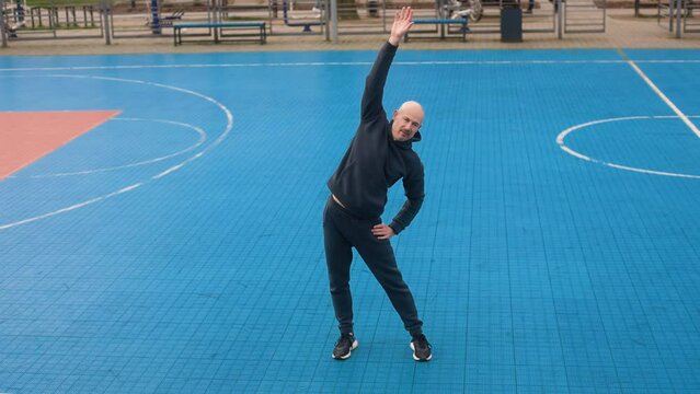A bald man in a sportswear is exercising on a city sports field. A male athlete is stretching on a city sports field. Full shot